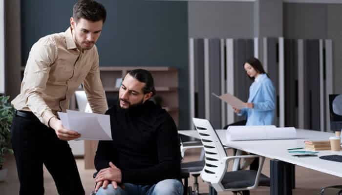 Employee morale can suffer after a serious incident | tycoonstory media Two colleagues discuss a document: one man standing and pointing to papers while the other sits and looks on in a modern office. In the background, a woman walks with a folder.