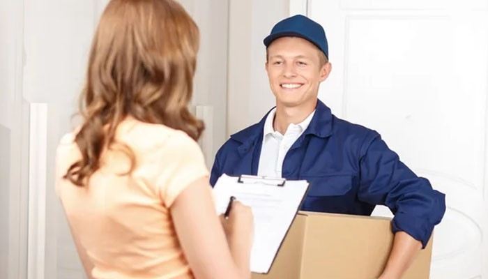 What happened after delivery | tycoonstory media Delivery person in a blue uniform hands a cardboard box to a woman at her doorway while she signs a clipboard, smiling.