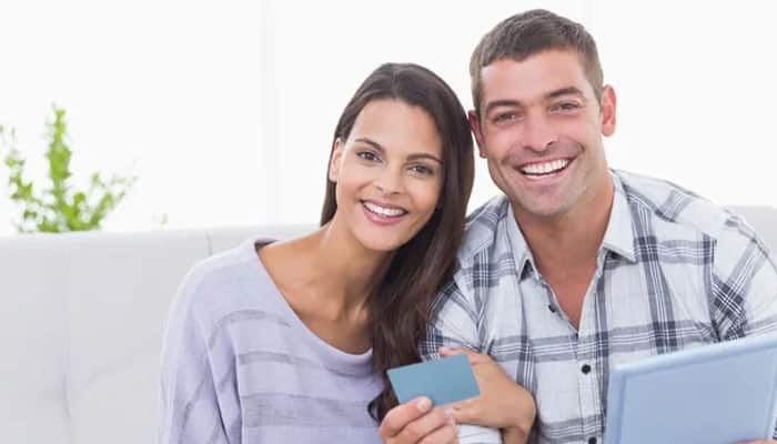 Securing your financial future with coast tradelines | tycoonstory media Smiling couple sitting on a light couch, the woman holding a blue card toward the camera and the man beside her with a laptop nearby.