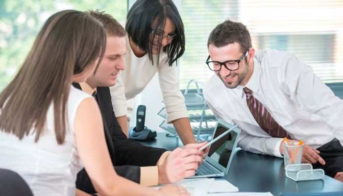 What it consulting can actually provide | tycoonstory media Four colleagues gathered around a laptop and tablet on a conference table, collaboratively looking at the screen in a bright office.