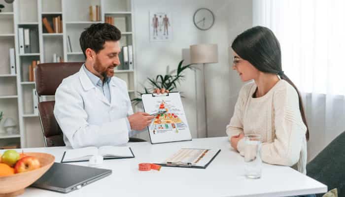 Sleep may be the closest thing we have to a mental health reset | tycoonstory media Male doctor in a white coat shows a colorful health/diet chart to a seated female patient at a desk in a bright clinic.