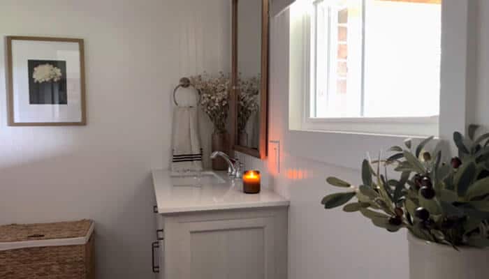 The new york city multiplier effect | tycoonstory media Cozy white bathroom vanity with a lit candle, vase, and dried flowers, near a window and a framed flower print on the wall.