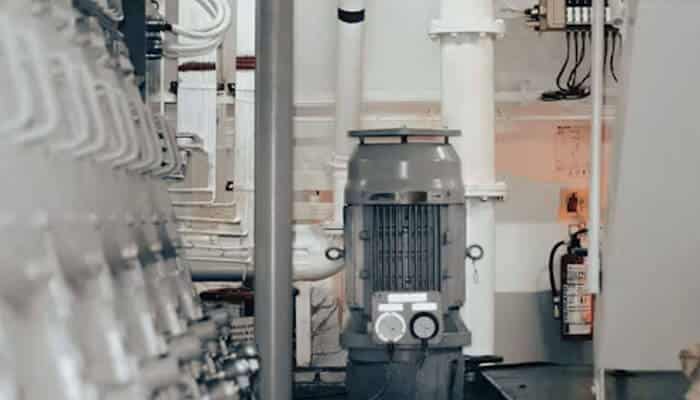 Why system evacuation comes before charging | tycoonstory media Gray vertical industrial pump with two gauges on the front, surrounded by white pipes in a mechanical room.