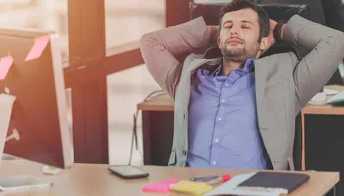 Use your body to steady yourself | tycoonstory media Man in a blue shirt and gray blazer leans back with hands behind head, eyes closed, at a cluttered desk in an office, appearing tired or relieved at work.