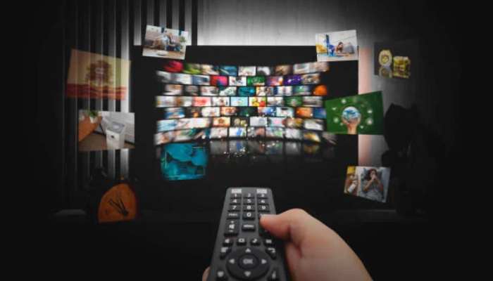 Hls vs mpeg-dash 1 | tycoonstory media Close-up of a hand holding a tv remote, pointed toward a wall-sized mosaic of colorful video thumbnails in a dark room, suggesting streaming content selection.