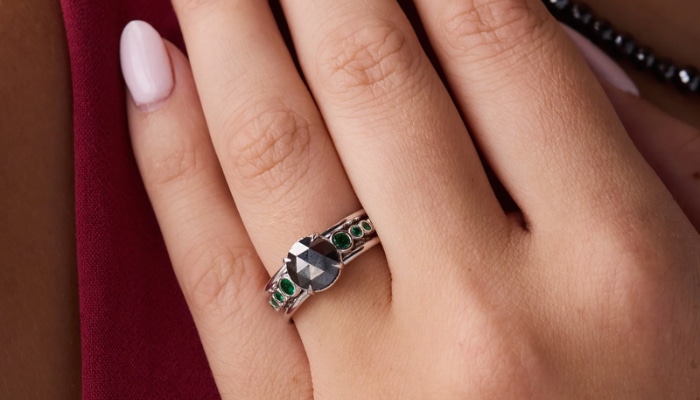 How a rose-cut black diamond engagement ring redefines style 1 | tycoonstory media Close-up of a woman's hand wearing a silver ring with a dark faceted central stone and small green stones along the band.