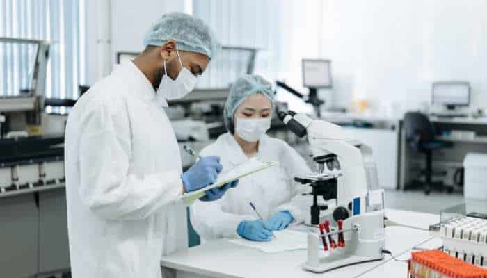 How tesamorelin interacts with ghrh receptors within experimental research models 3 | tycoonstory media Two researchers in lab coats and masks review notes beside a microscope at a lab bench in a bright lab environment.