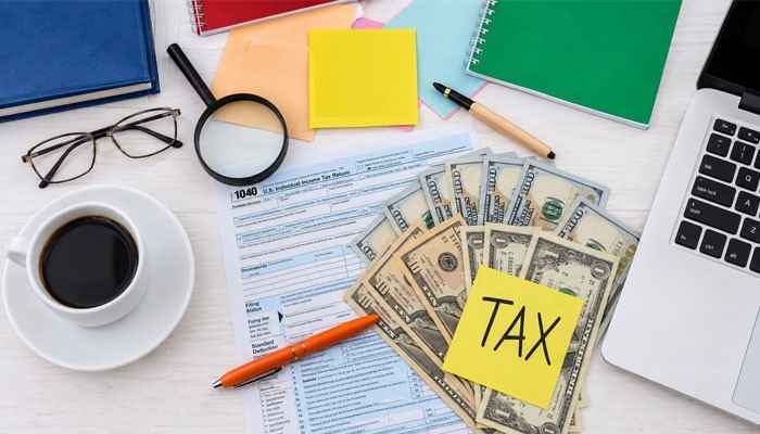 The 7 best services for catching up overdue tax returns in australia | tycoonstory media Desk with tax forms, cash, a yellow 'tax' sticky note, laptop, coffee, and glasses.