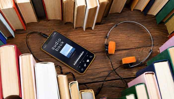 Audio arrives with the picture | tycoonstory media Smartphone playing an audiobook surrounded by colorful books and orange headphones on a wooden surface.