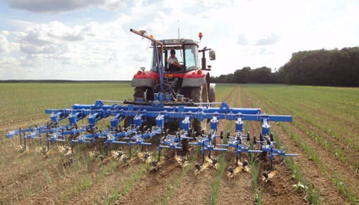 Picture shown:ptx trimble autopilot steering system | tycoonstory media Red tractor pulling a blue cultivator across a field, hoeing rows of young crops under a cloudy sky.
