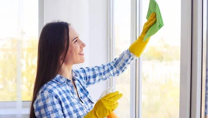 Window tracks and frames collect more than dust | tycoonstory media Smiling woman cleaning a glass window with a green cloth, wearing yellow rubber gloves and a blue plaid shirt, holding a spray bottle.