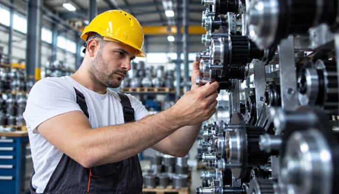 Mechanical failures and the maintenance chain | tycoonstory media Factory worker in a yellow hard hat adjusts a large metal gears assembly in an industrial workshop, focusing on the machinery.