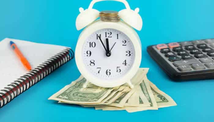 The 7 mistakes that cost time and money | tycoonstory media White alarm clock sitting on a fan of dollar bills, with a notebook and calculator on a bright blue background (time is money).
