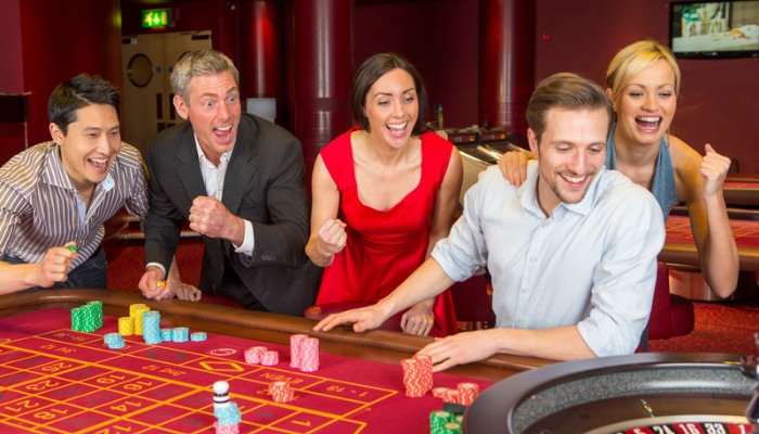 Online casino platforms 1 | tycoonstory media A group of five adults cheers at a casino roulette table with colorful chips and a smiling dealer-like player in front.