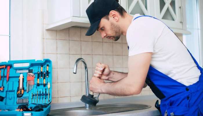 Roto-rooter services_ expert guide for homeowners 1 | tycoonstory media Plumber repairing a kitchen sink faucet, using a wrench beside an open tool kit on the counter.
