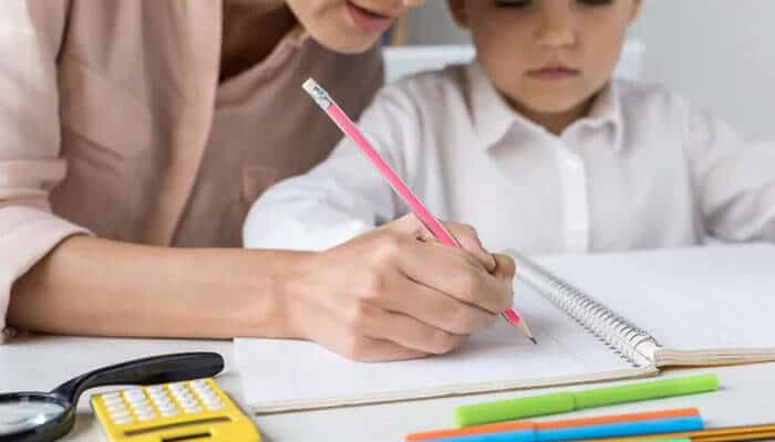 Review the plan as your child grows | tycoonstory media Adult helping a child write with a pink pencil at a white desk, surrounded by a notebook, calculator, ruler and colorful markers.