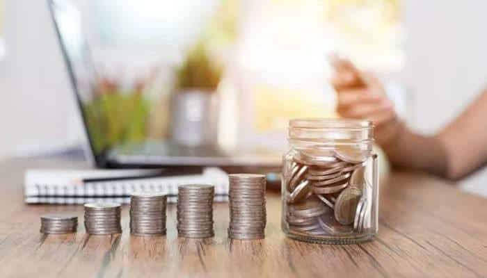 The four bucket method for building your budget | tycoonstory media Coin stacks of increasing height on a wooden desk with a jar of coins nearby and a laptop in the blurred background.