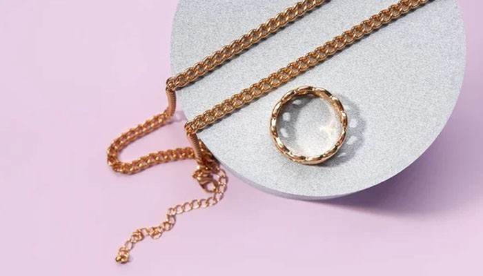 Watch what changes around the hoop | tycoonstory media Rose gold necklace with a matching ring draped over a gray display circle on a pastel pink background.