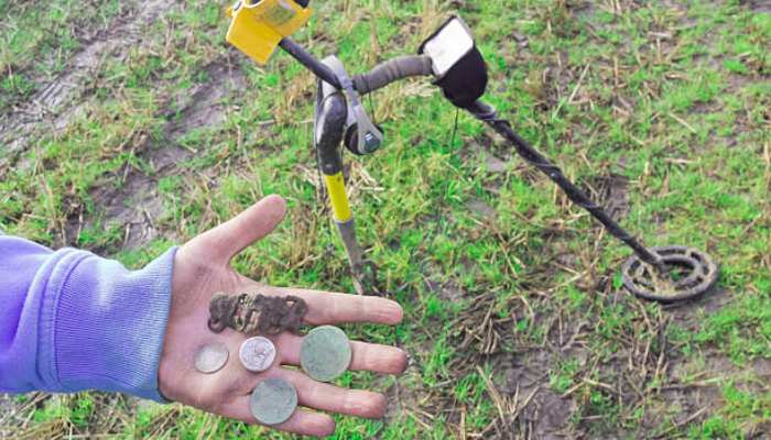 What do metal detectorists find most often 1 | tycoonstory media Person's gloved hand displays various coins and metal finds after using a metal detector in a grassy, muddy field with the detector in the background.