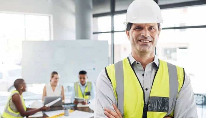 Protecting employees builds stronger businesses | tycoonstory media Construction professional in a safety vest and hard hat stands with arms crossed, smiling in a meeting room with colleagues in the background