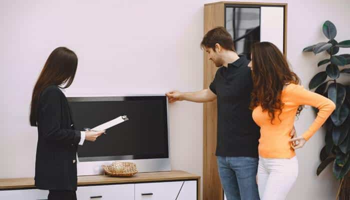Choose the size in the room, not on a screen | tycoonstory media Three people in a living room look at a flat-screen tv on a cabinet; the woman in black holds papers while the other two discuss nearby.