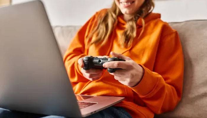Choose an eligible game pass plan - tycoonstory | tycoonstory media Woman in an orange hoodie using a controller and laptop for xbox cloud gaming, enjoying console-free gaming from the comfort of her home.