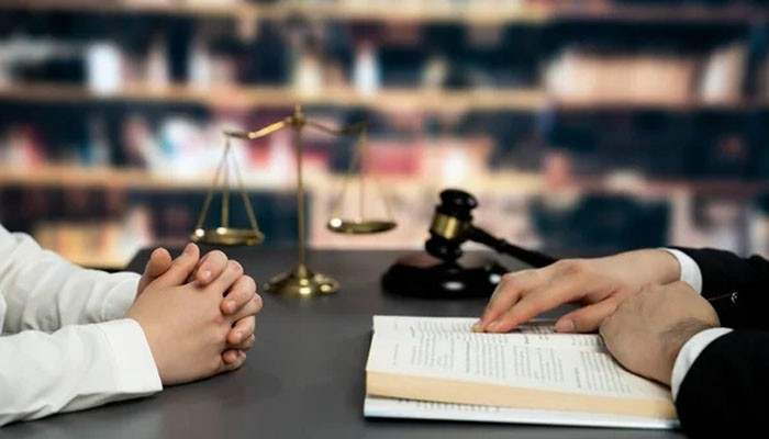 Avard law offices | tycoonstory media Hands clasped at a desk with scales of justice and a gavel in the background, next to an open book, suggesting a legal setting.