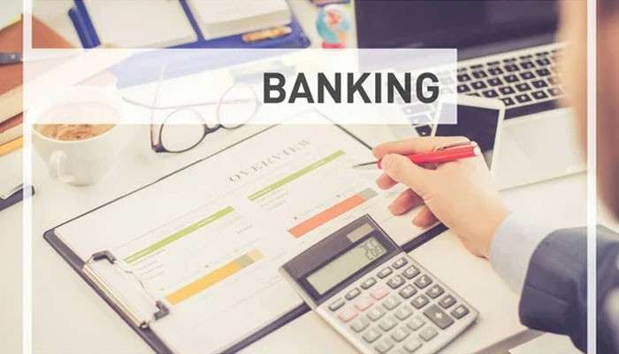 How does a business bank account fees comparison work in practice? | tycoonstory media Person writes on banking documents with a calculator and laptop on a desk, a pair of glasses nearby, overlaid with the word banking.