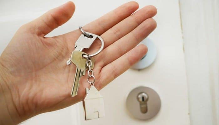 If you're shopping for a rental property loan | tycoonstory media Hand holding a keyring with house-shaped charm in front of a door lock, suggesting moving in or home access.
