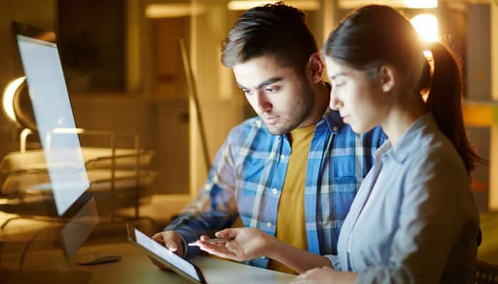 Top 10 enterprise software & app testing companies | tycoonstory media Two coworkers collaborate at a desk, focusing on a tablet in front of a monitor under warm office lighting.