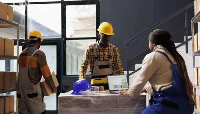 Prepare the information and the delivery team | tycoonstory media Three construction workers in yellow hard hats collaborate in a warehouse; a woman in blue overalls uses a laptop at a table with a blue helmet nearby.