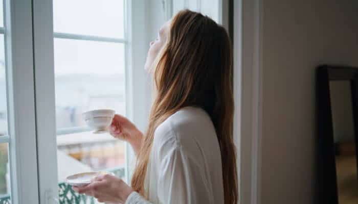 Decorating for your unique lifestyle needs | tycoonstory media Woman in a white robe stands by a window, holding a teacup and saucer, looking upward toward the light.