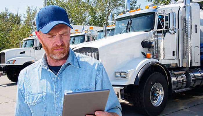 The truck's electronic records may matter | tycoonstory media Male truck driver in a blue cap and denim shirt reads a tablet beside a line of white semi-trucks in a truck yard.