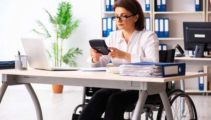 Permanent disability payments | tycoonstory media Woman in a wheelchair at an office desk, using a calculator with a laptop nearby.