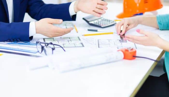 Apply for the nj compliance bond | tycoonstory media Team of engineers reviewing blueprints on a table with a calculator, glasses, pencils and an orange hard hat.