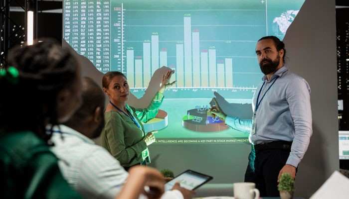 | tycoonstory media Two colleagues present analytics to a group, pointing at a large screen filled with bar charts in a modern conference room.
