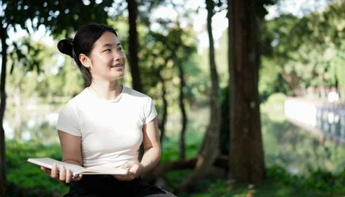 Learning to sit with silence | tycoonstory media Young woman sitting in a park, holding a book and looking to the side with a gentle smile, surrounded by trees.