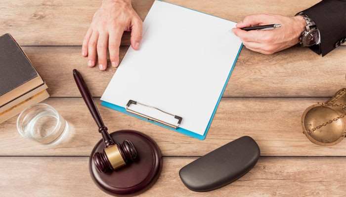 5280 injury law | tycoonstory media Top-down view of a wooden desk with a gavel, clipboard holding a blank sheet, a hand writing with a pen, and a small heart-shaped dish nearby.