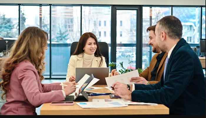 Build hiring lead time into the forecast | tycoonstory media Four professionals sit around a conference table in a bright office, reviewing papers and a laptop together.