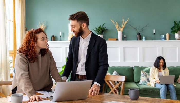 Making compromise part of everyday business and relationship life | tycoonstory media Two colleagues stand at a wooden table with laptops, talking and smiling, while a person on a green couch works on a laptop in the background in a bright office.