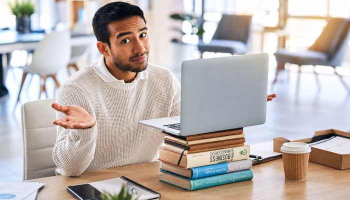 Choose an all-in-one platform | tycoonstory media Man in a white sweater sits at a desk with a laptop resting on stacked books, shrugging his shoulders.
