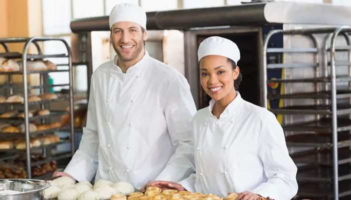 Reduce repetitive staff work | tycoonstory media Two bakers in white uniforms and hats stand behind a counter with trays of pastries, smiling at the camera in a bakery.