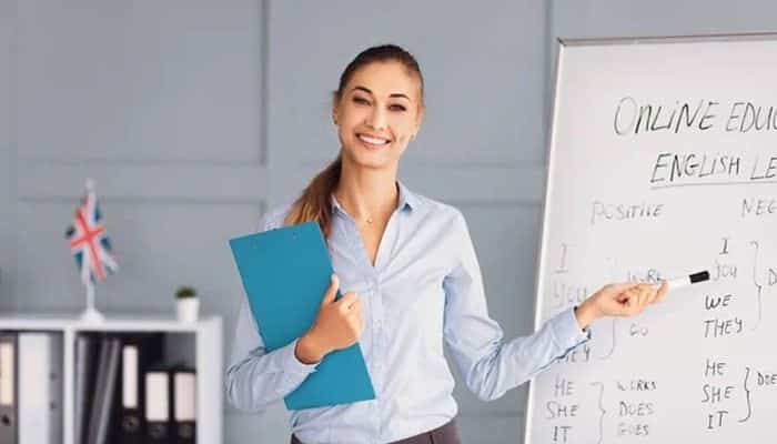 The link between language and career progression | tycoonstory media Smiling female teacher in a light blue shirt stands beside a whiteboard, holding a blue folder during an english lesson.