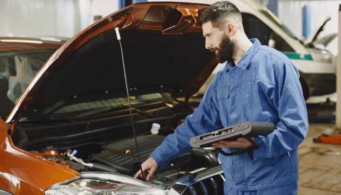 What is the history and condition of this car? | tycoonstory media Male mechanic in blue coveralls inspecting a car engine under an open hood, holding a diagnostic device.