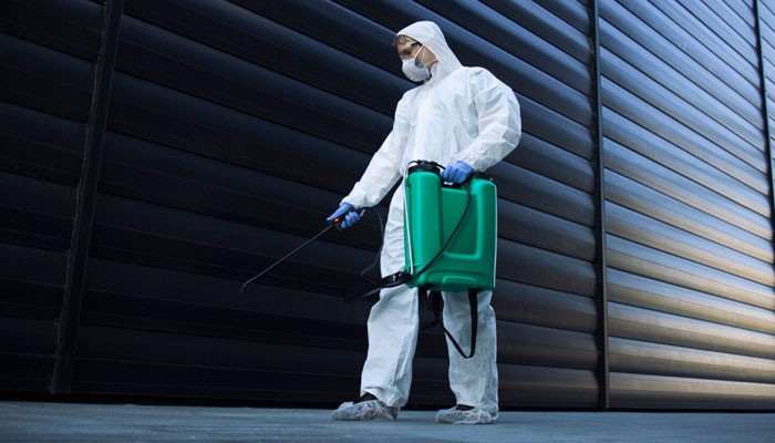 Study local demand and service gaps | tycoonstory media Worker in full protective hazmat suit and mask wearing a green backpack sprayer, disinfecting an industrial metal wall exterior.