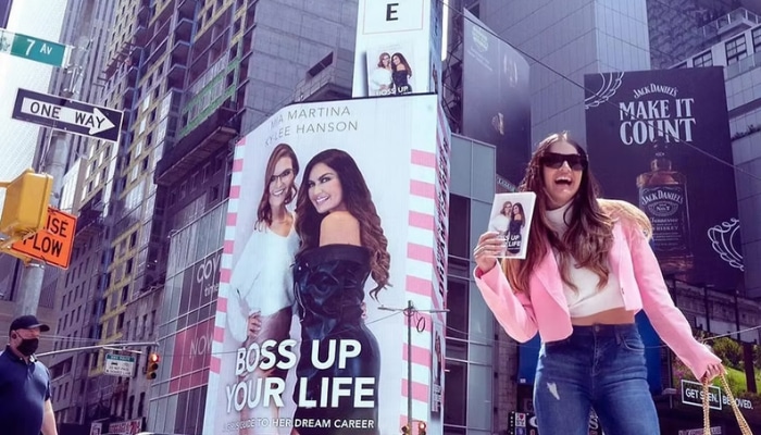Plug talk media 1 | tycoonstory media Smiling woman in a pink jacket poses with a phone in times square, large 'boss up your life' billboard behind her.
