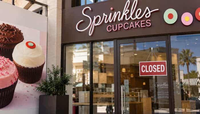 Why did sprinkles cupcakes close - tycoonstory | tycoonstory media Sprinkles cupcakes storefront with a closed sign on the glass entrance and large cupcake images displayed beside the bakery.