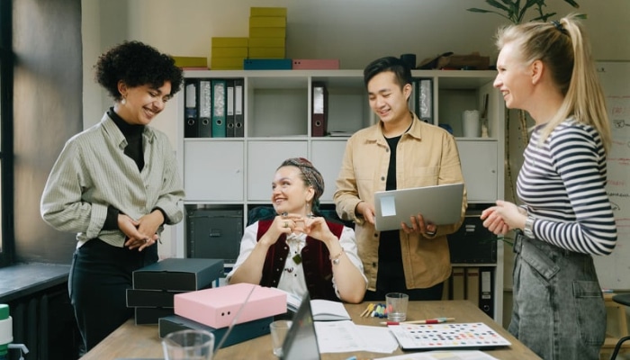 Why traditional workplace perks fail growing companies and how to fix them 1 | tycoonstory media Four coworkers in a bright office stand and sit around a desk, with a seated woman showing a mug as colleagues smile and chat; a man holds a laptop nearby.
