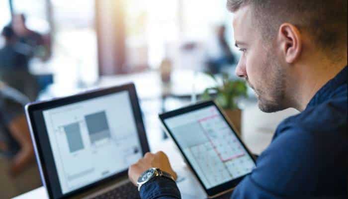 Product configuration and version control | tycoonstory media Man in a blue shirt works on a laptop and a tablet at a bright desk, reviewing charts on both screens.