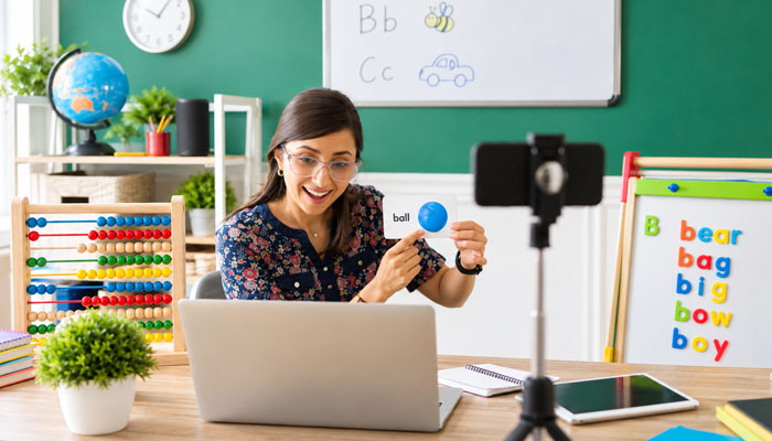 Which platform is a better fit for you | tycoonstory media Teacher records a phonics lesson for kids, holding a blue ball and a card that says'ball' in a bright classroom setup with a laptop and teaching tools nearby.